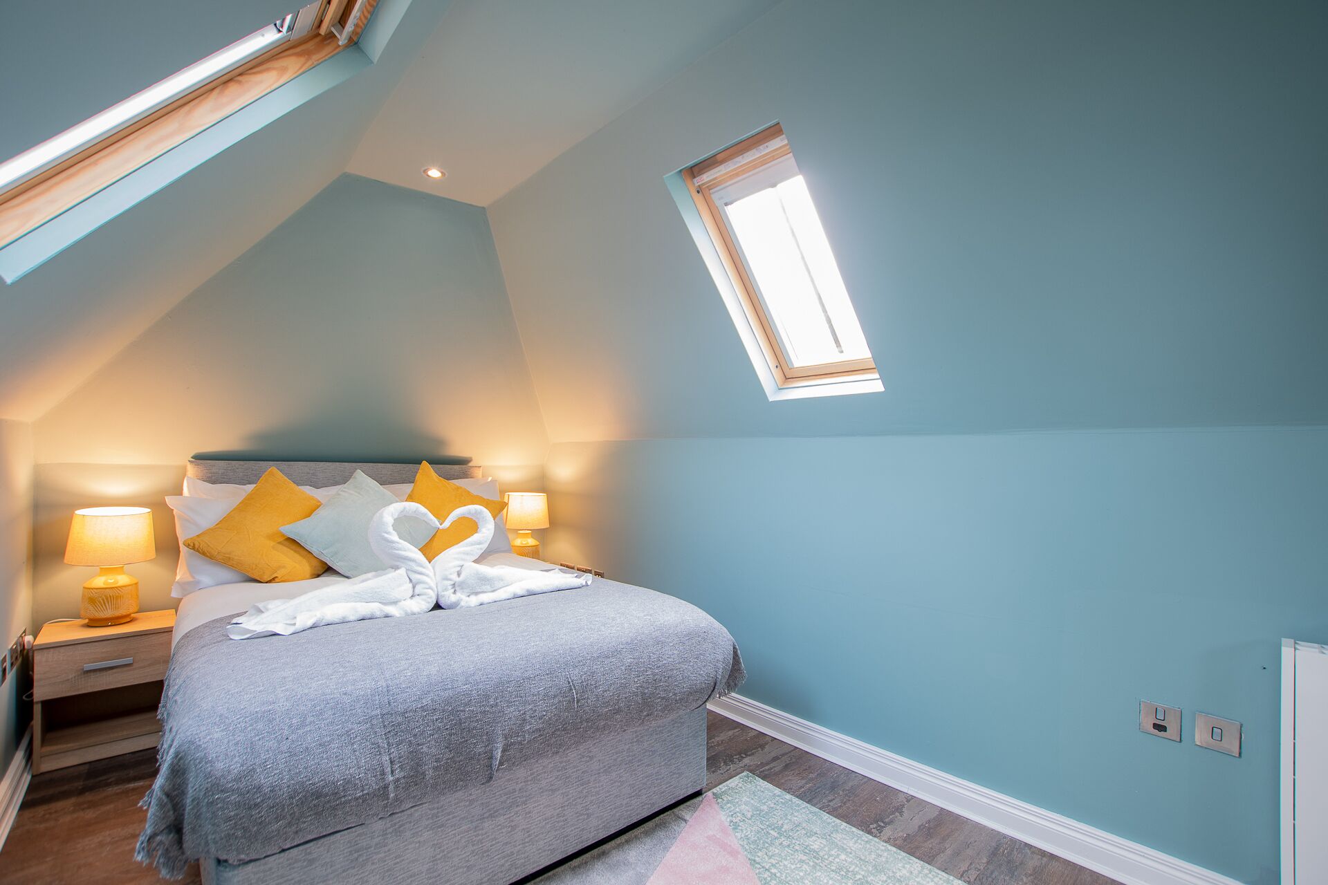 A bedroom with a sloped ceiling, a skylight, a bed with a grey blanket, two bedside lamps, and a heart-shaped towel arrangement.