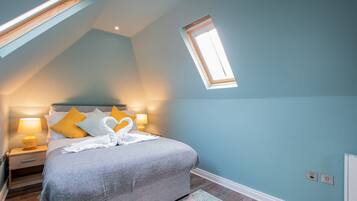 A bedroom with a sloped ceiling, a skylight, a bed with a grey blanket, two bedside lamps, and a heart-shaped towel arrangement.