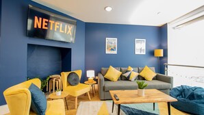 Un salon avec un téléviseur à écran plat affichant le logo « NETFLIX », un canapé avec des coussins, un fauteuil jaune, une table basse en bois et un mur bleu.
