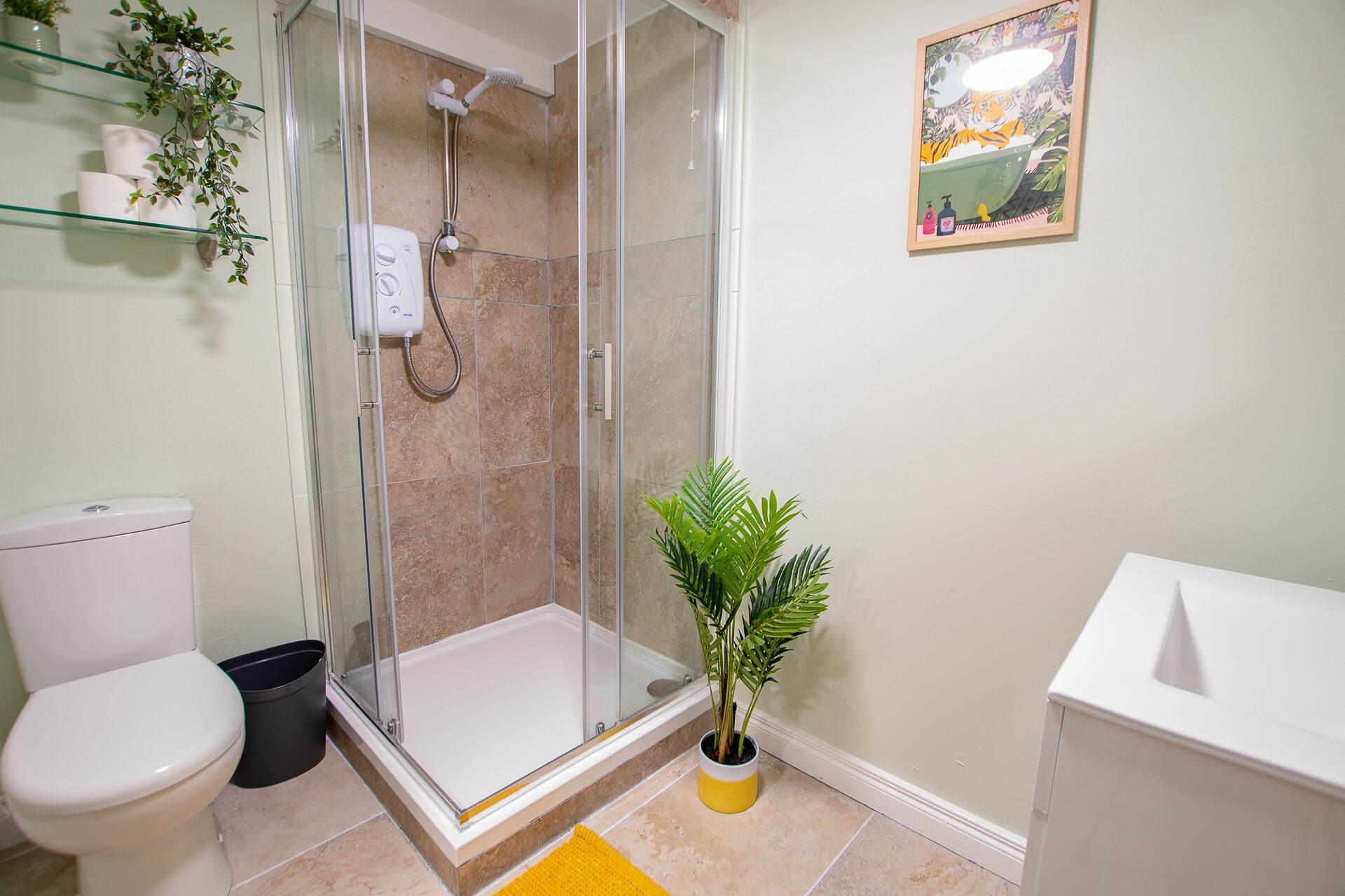 A bathroom with a glass-enclosed shower, a toilet, a trash bin, a sink, and a potted plant.