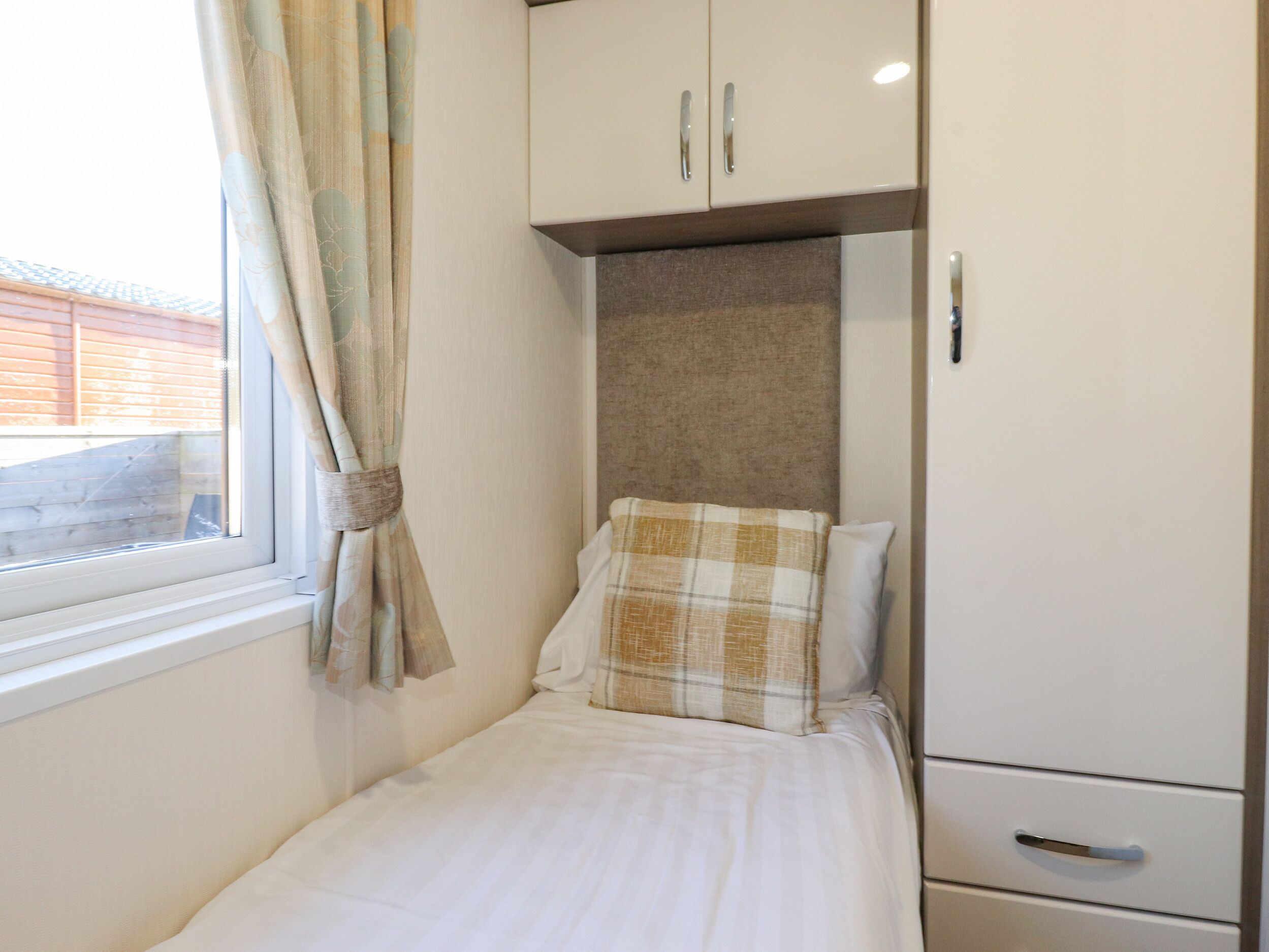 A small, neatly made bed with a white sheet and a plaid pillow, a window with curtains, and a white wardrobe with two doors.