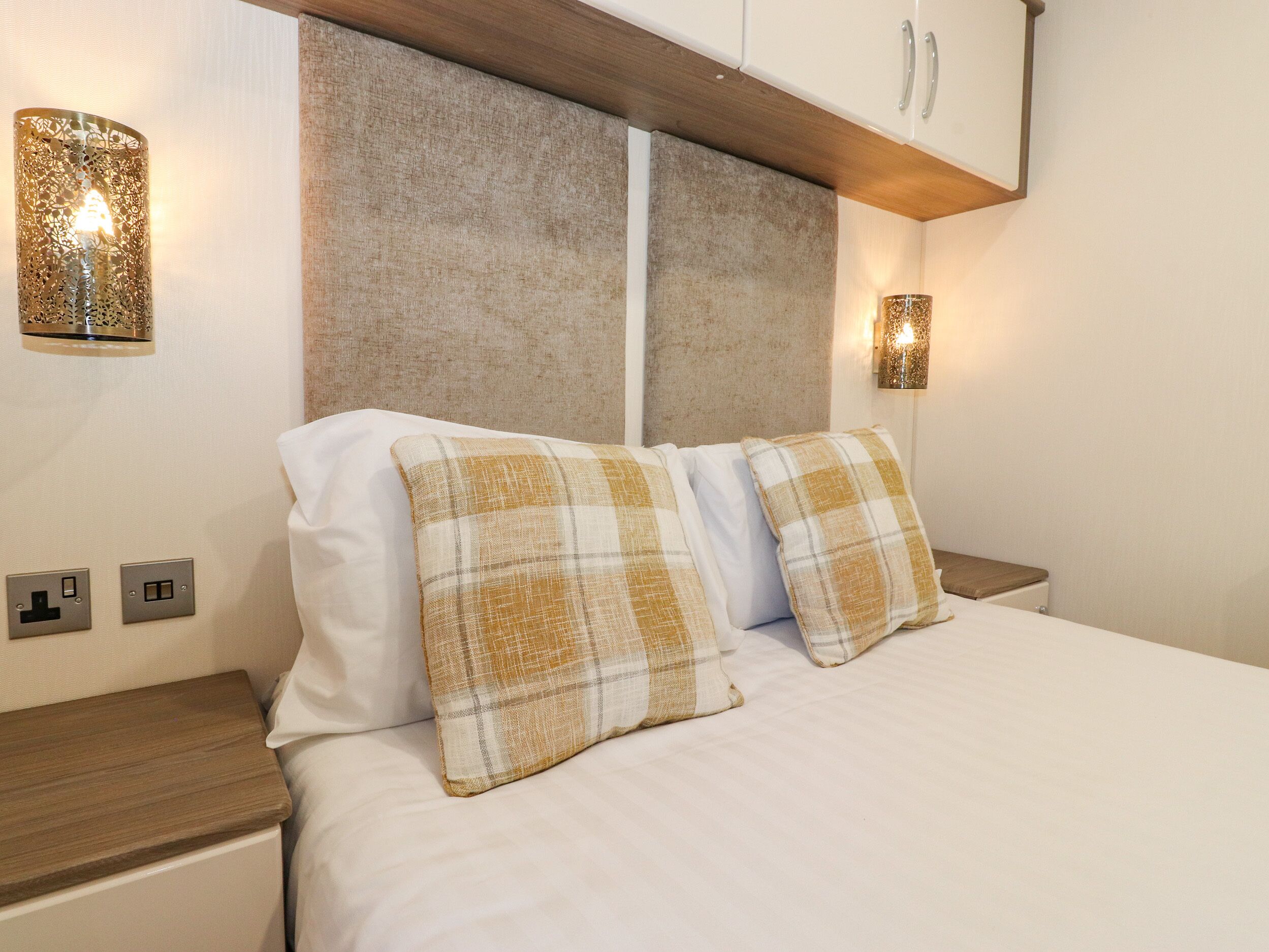 A neatly made bed with two plaid pillows, a headboard with a textured finish, and two wall-mounted lamps on either side.