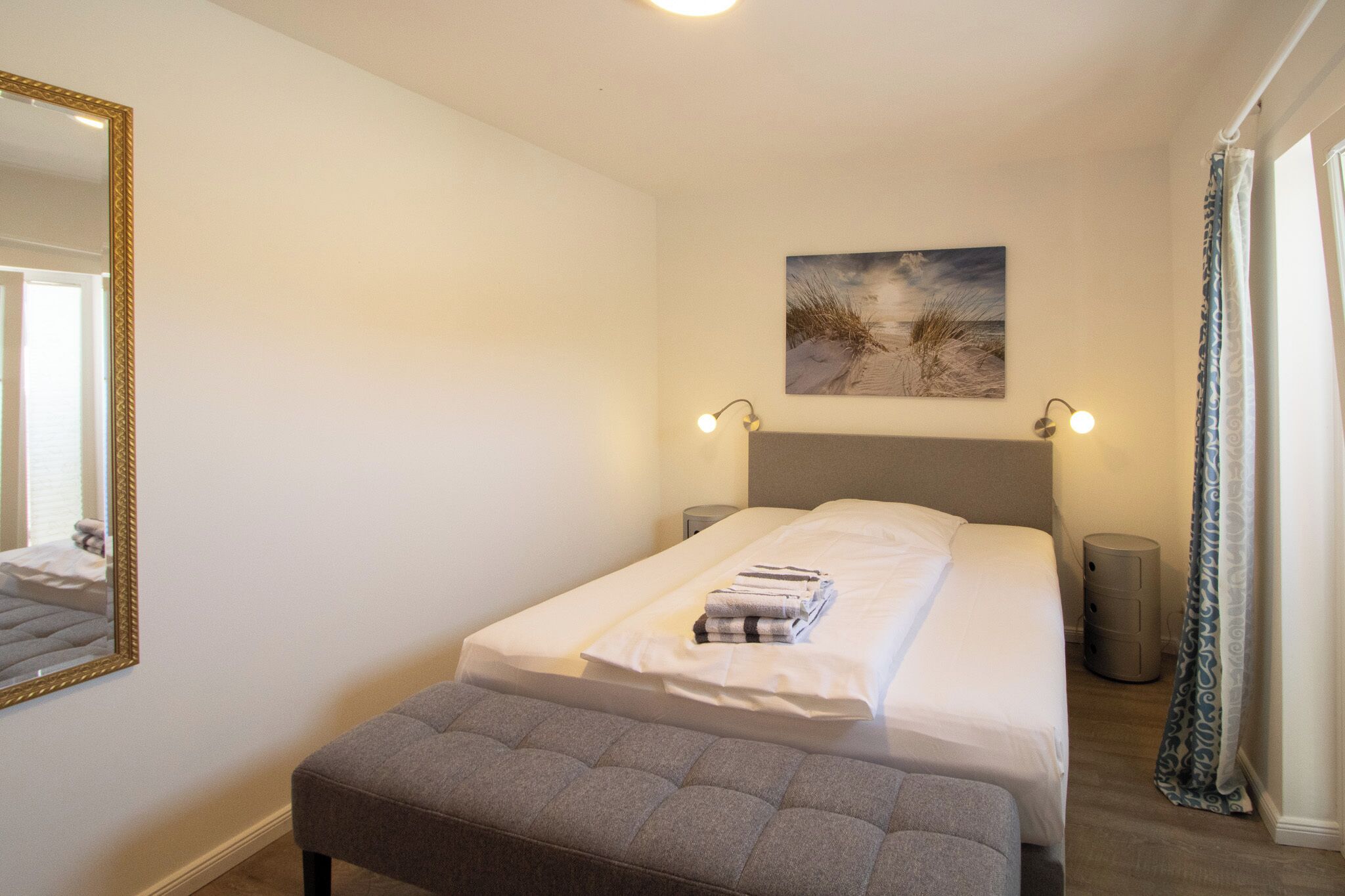 Cama bem arrumada com cabeceira, abajur, quadro pendurado na parede, espelho e janela com cortinas.