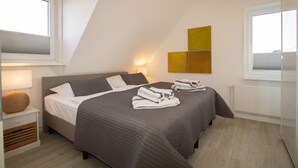 Quarto com duas camas, uma mesa de cabeceira com abajur, iluminação na parede e janela com persianas.