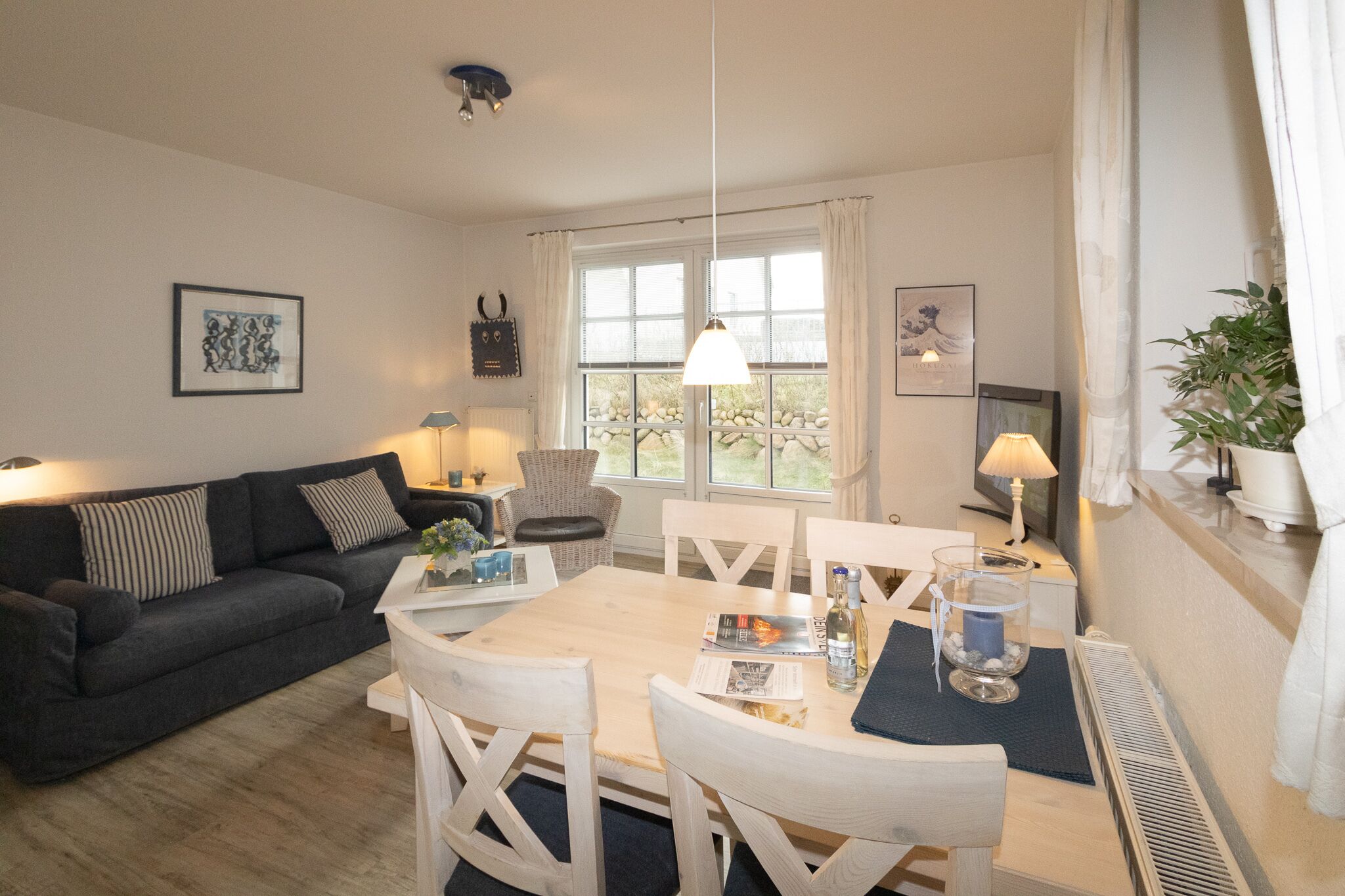 Un salon chaleureux avec un canapé gris foncé, une table à manger en bois et quatre chaises, ainsi qu’une grande fenêtre agrémentée de rideaux.