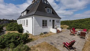 Ein weißes Haus mit einem steilen Dach, einer kleinen Veranda mit roten Stühlen und einer gepflasterten Terrasse.