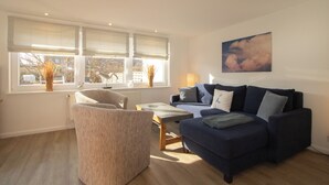 Un salon avec un canapé bleu, un fauteuil beige, une petite table et une fenêtre avec des stores.