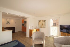 Un salon moderne avec un téléviseur à écran plat, un canapé et un fauteuil. On aperçoit au fond une cuisine avec des meubles en bois et un coin repas.