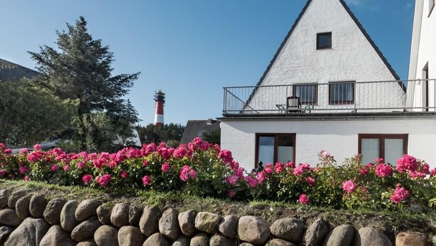 Un bâtiment blanc avec un toit triangulaire, un balcon avec des chaises et des fleurs roses au premier plan.