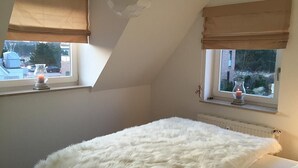 Quarto com cama fofa branca, duas janelas com cortinas bege e um radiador.