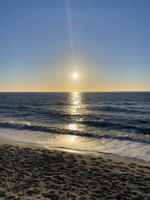 A beach with footprints leading to the water, waves gently crashing onto the shore, and the sun reflecting off the water.