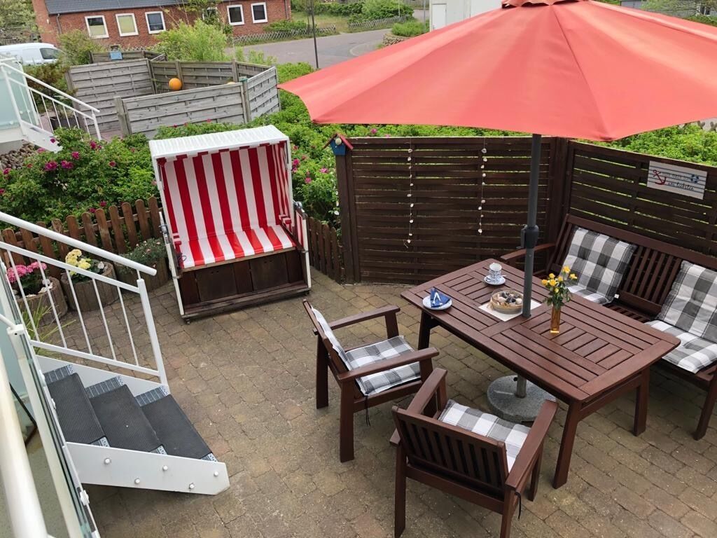 A patio area with a wooden table, two chairs, a striped bench, and a red umbrella.