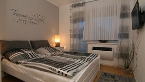 Ett sovrum med en säng, ett nattduksbord med en lampa, en tv som är monterad på väggen och ett fönster med tunna, genomskinliga gardiner.