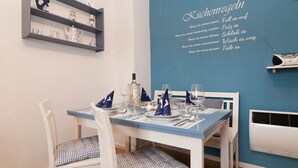 En matplats med en blå vägg, ett vitt bord och stolar med rutiga kuddar. Det finns hyllor med dekorativa föremål och en väggmonterad skylt med text.