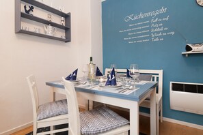 Un comedor con pared azul, mesa blanca y sillas con cojines a cuadros. Hay estantes con objetos decorativos y un letrero en la pared.