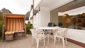 A patio with a white table and chairs, a striped bench, and a view of a house through a window. - Vacation apartment for up to 4 people (2 adults + 2 children) (Hörnum auf Sylt)