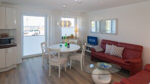 A modern living room with a red sofa, a glass coffee table, a dining table with chairs, a TV, and a kitchen area with a microwave. - Directly on the beach with a view of the sea (Hörnum auf Sylt)