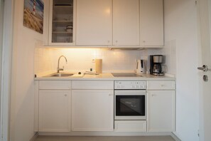 Cocina compacta con muebles blancos, horno empotrado, cafetera y lavamanos.