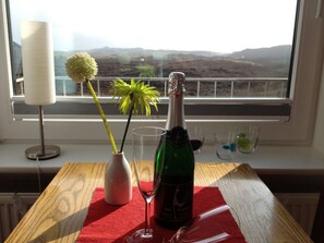 Une table avec une bouteille de vin, un verre, un vase avec une fleur et une bougie, le tout posé sur un tapis rouge près d’une fenêtre donnant sur des collines.