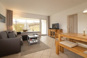 A modern living room with a sofa, coffee table, and a dining area with a wooden table and benches. - Attractive vacation apartment in an absolutely quiet location in the Hörnum dunes (Hörnum auf Sylt)