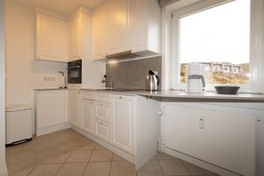 A modern kitchen with white cabinetry, a built-in oven, and a window with a view of buildings. - Attractive vacation apartment in an absolutely quiet location in the Hörnum dunes (Hörnum auf Sylt)