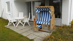 Un petit espace extérieur aménagé avec des sièges, comprenant une chaise à rayures et deux chaises blanches, une petite table et une fenêtre avec des rideaux.