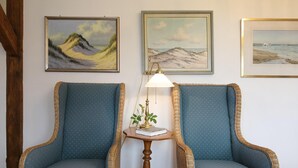Two wicker armchairs with blue upholstery, a small wooden table with a lamp, and three framed paintings on the wall.