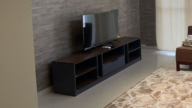 A modern living room with a flat-screen TV on a dark wooden stand, a wall-mounted air conditioner, and a large window with curtains.