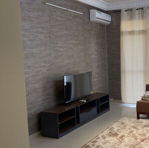 A modern living room with a flat-screen TV on a dark wooden stand, a wall-mounted air conditioner, and a large window with curtains.
