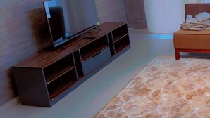 A modern living room with a flat-screen TV on a wooden stand, a bed with a headboard, and a patterned rug.