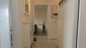 A narrow hallway with white walls and a tiled floor, leading to a room with a bed and a window.