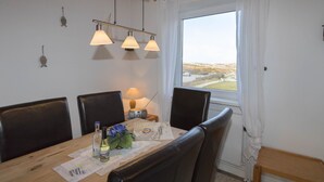 A dining area with a wooden table, four chairs, a radio, and a window with a view of the countryside. - Cozy vacation apartment in the immediate vicinity of.... (Hörnum auf Sylt)
