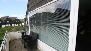 A balcony with a table and chairs, a large window with blinds, and a view of neighboring houses. - Cozy vacation apartment in the immediate vicinity of.... (Hörnum auf Sylt)