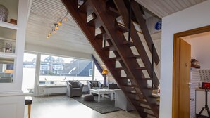 A spacious living area with a wooden staircase, a sofa set, a coffee table, and a view of buildings outside. - Cozy vacation apartment in the immediate vicinity of.... (Hörnum auf Sylt)