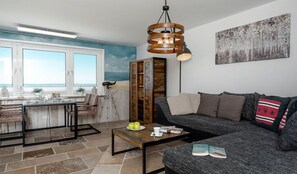 Ein Wohnzimmer mit Blick auf den Ozean, einem Holztisch, einer Couch mit Kissen und einem Gemälde an der Wand.