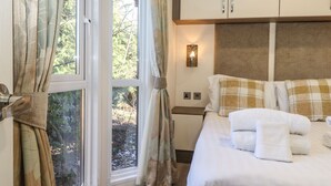 A hotel room with a bed, curtains, a nightstand, a wall-mounted lamp, and a window with a view of trees. - Hercules (Morpeth)