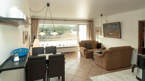 A modern living room with a sofa, dining table, and chairs. There is a large window with a view of the outdoors. - Attractive vacation apartment in an absolutely quiet location in the Hörnum dunes (Hörnum auf Sylt)