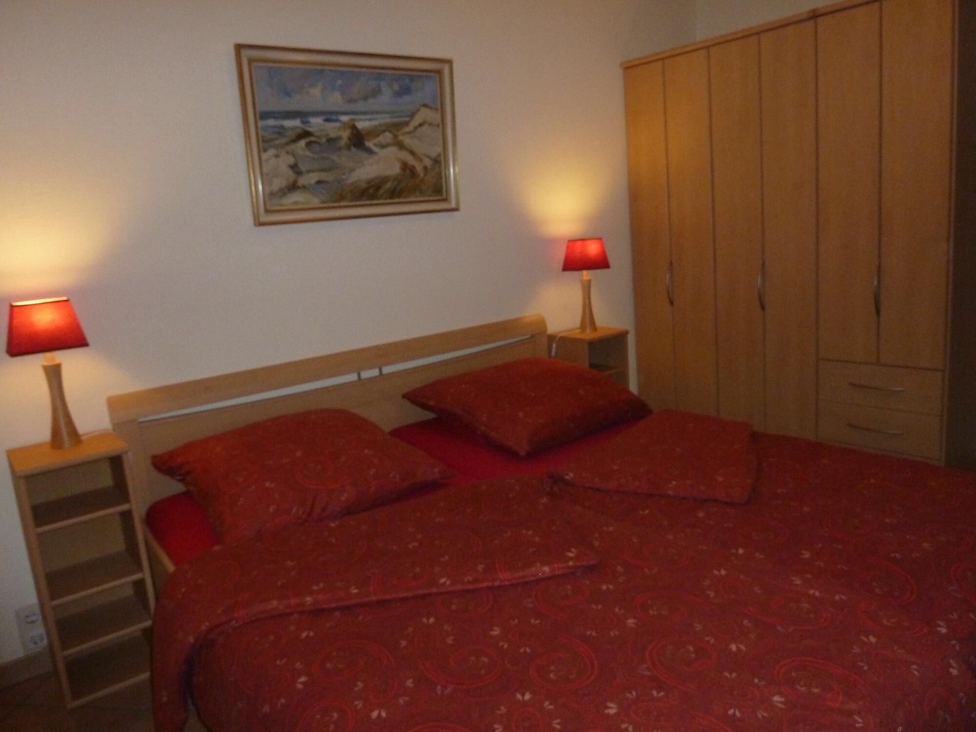 A bedroom with a wooden bed, red bedding, two bedside lamps, a wooden wardrobe, and a painting on the wall.