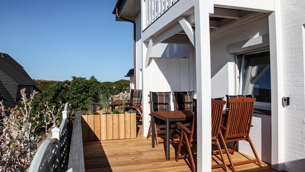 A wooden deck with a table and chairs, a white building with a balcony, and a clear blue sky. - The Leuchtfeuer apartment is located in the Atair house in Hörnum.... (Hörnum auf Sylt)