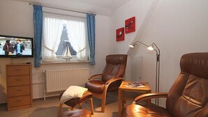 Une pièce avec une télévision, une fenêtre avec des rideaux, un radiateur, un fauteuil en cuir marron, une table en bois et une lampe.