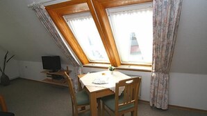 Una habitación con techo inclinado, una mesa de comedor con sillas, un televisor sobre un mueble bajo y una ventana con cortinas.