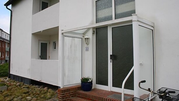 Un edificio de dos pisos con fachada blanca, un balcón en el nivel superior y una entrada con puerta de vidrio corredera.