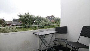 Un balcón con una mesa metálica y dos sillas, una pared blanca y vistas a casas y vegetación.