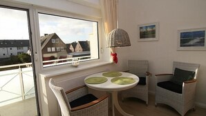 Een kamer met rieten stoelen, een ronde tafel met groene tafelonderleggers, en een uitzicht op huizen door een groot raam.