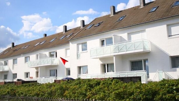 Een rij witte appartementen met balkons, een rode vlag en een heldere blauwe lucht.