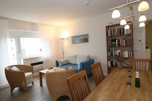 A cozy living room with a blue sofa, wicker chairs, a wooden dining table, and a bookshelf filled with books.