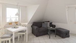 Ein stilvolles, weißes Zimmer mit einer grauen Couch, einem kleinen runden Tisch und einem weißen Esstisch mit Stühlen.