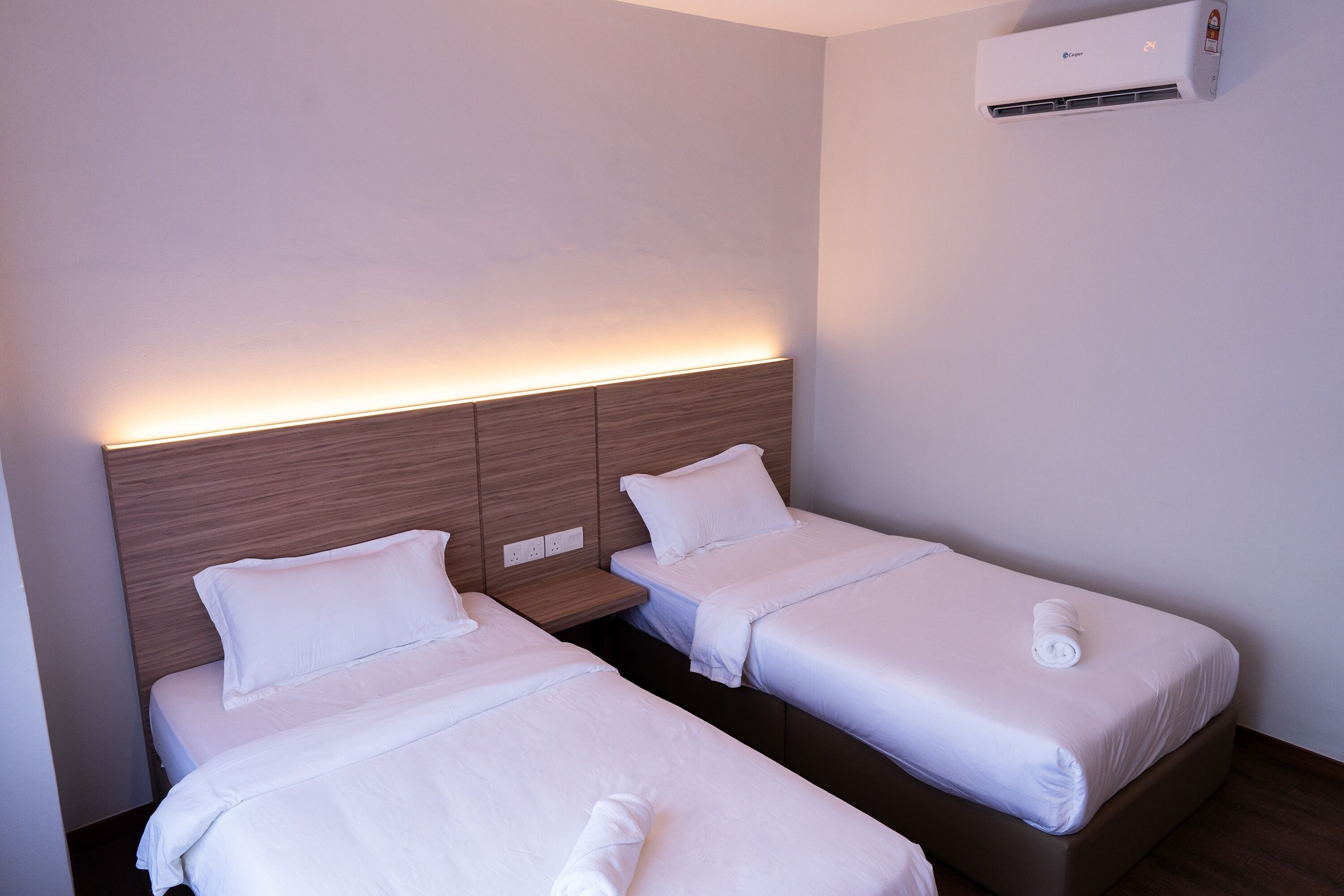Two single beds with wooden headboards and white bedding, a wall-mounted air conditioner, and a small bedside table with a lamp.