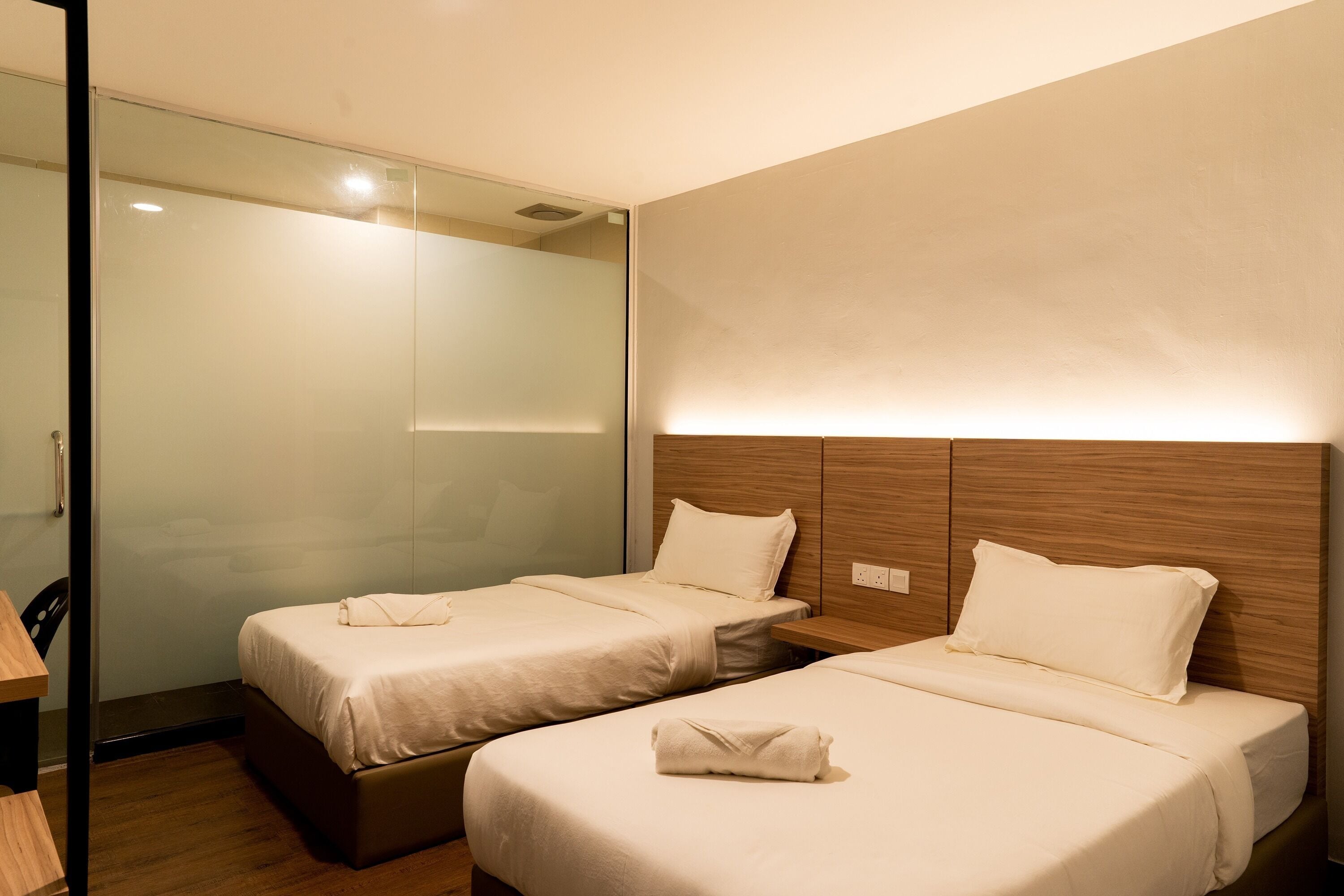 A hotel room with two beds, a wooden headboard, a glass partition, and a wooden desk.