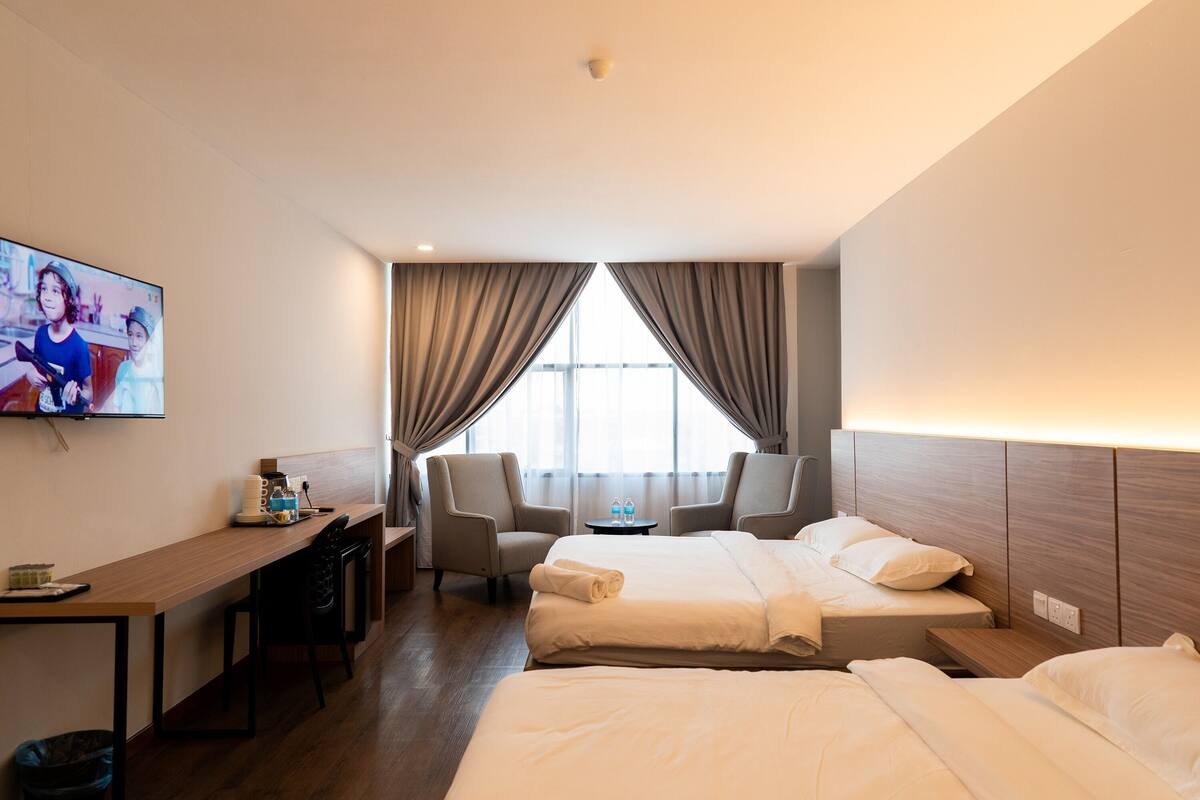 Ein Hotelzimmer mit einem Bett, einem Schreibtisch, zwei Stühlen, einem Fernseher und einem Fenster mit Vorhängen.